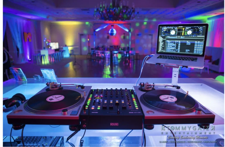 DJ setup with turntables and a laptop in a colorful, dimly lit event space. Tables and chairs are in the background under a chandelier.
