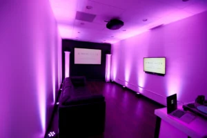 A room with purple lighting, featuring a dark sofa, a wall-mounted TV, and a projector screen. The room has a modern, minimalistic design.