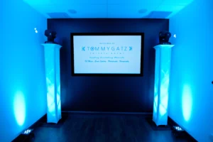 A sign reads "Weddings by Tommy Gatz Entertainment." Two illuminated pillars with lights flank a screen on a dark wall in a room with a blue glow.