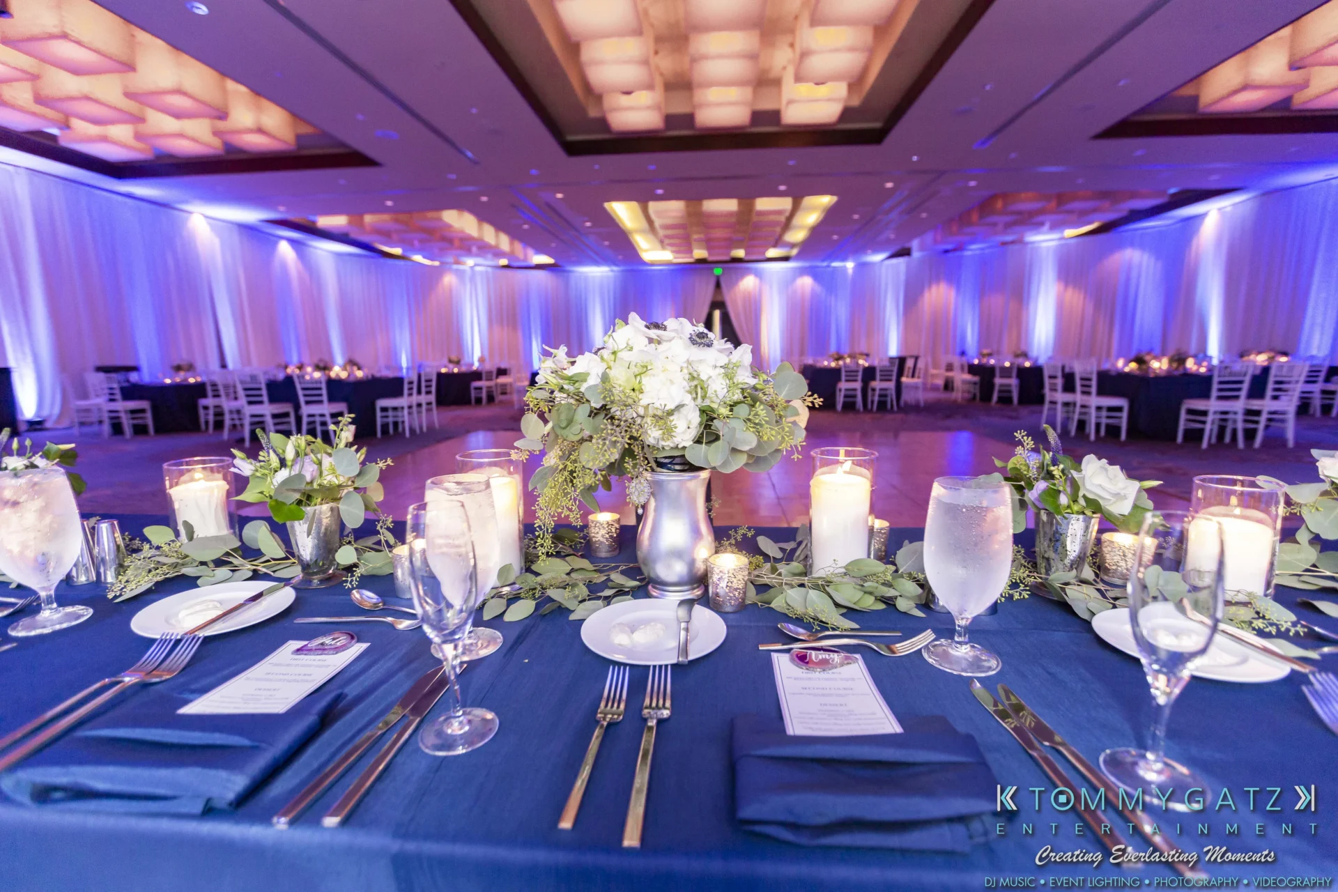 Elegant wedding reception hall with blue and purple lighting. A table is set with blue cloth, white floral centerpiece, candles, glassware, and cutlery.