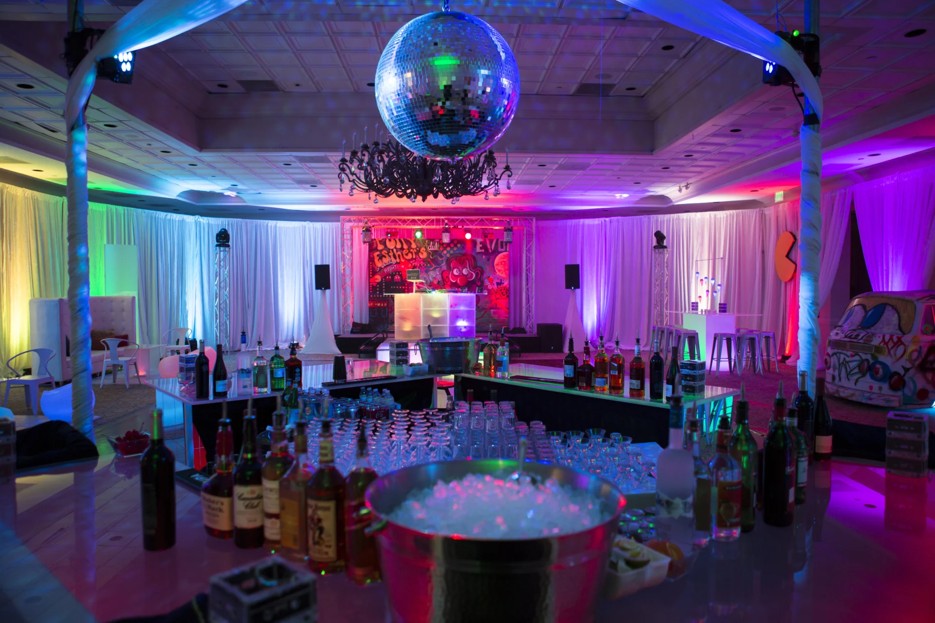 A colorful party bar setup with bottles, a large ice bucket, and a disco ball overhead. Stage with DJ equipment in the background, surrounded by vibrant drapes and atmospheric lighting.