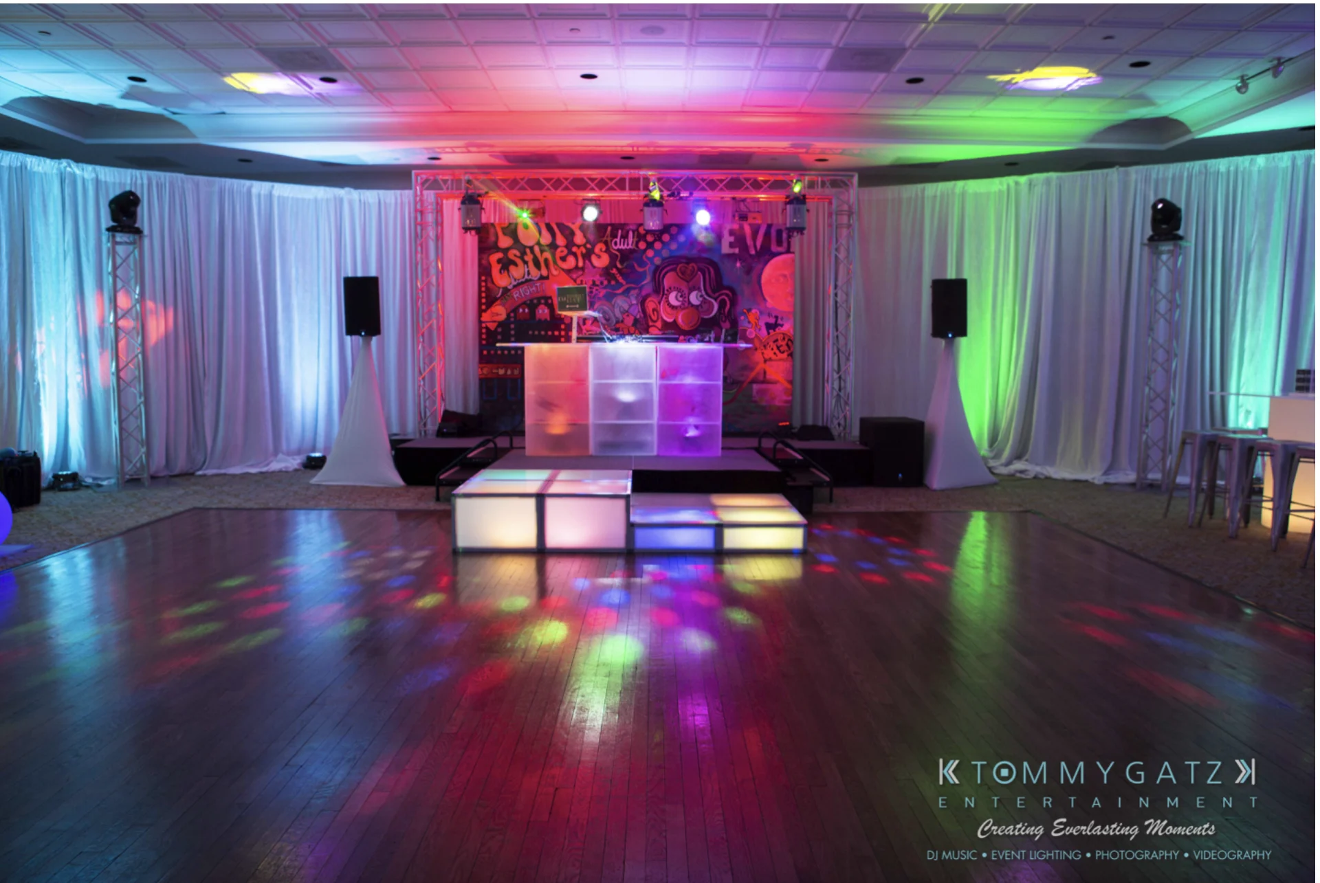 A dance floor with colorful lights and DJ booth, surrounded by speakers and white curtains. Bright graffiti mural in the background.