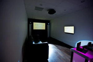 A dark room with a couch faces two screens displaying "Tommy Gatz." The room has wood flooring and overhead lighting.
