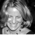 Black and white photo of a woman with shoulder-length hair smiling while looking down, wearing a necklace and earrings.