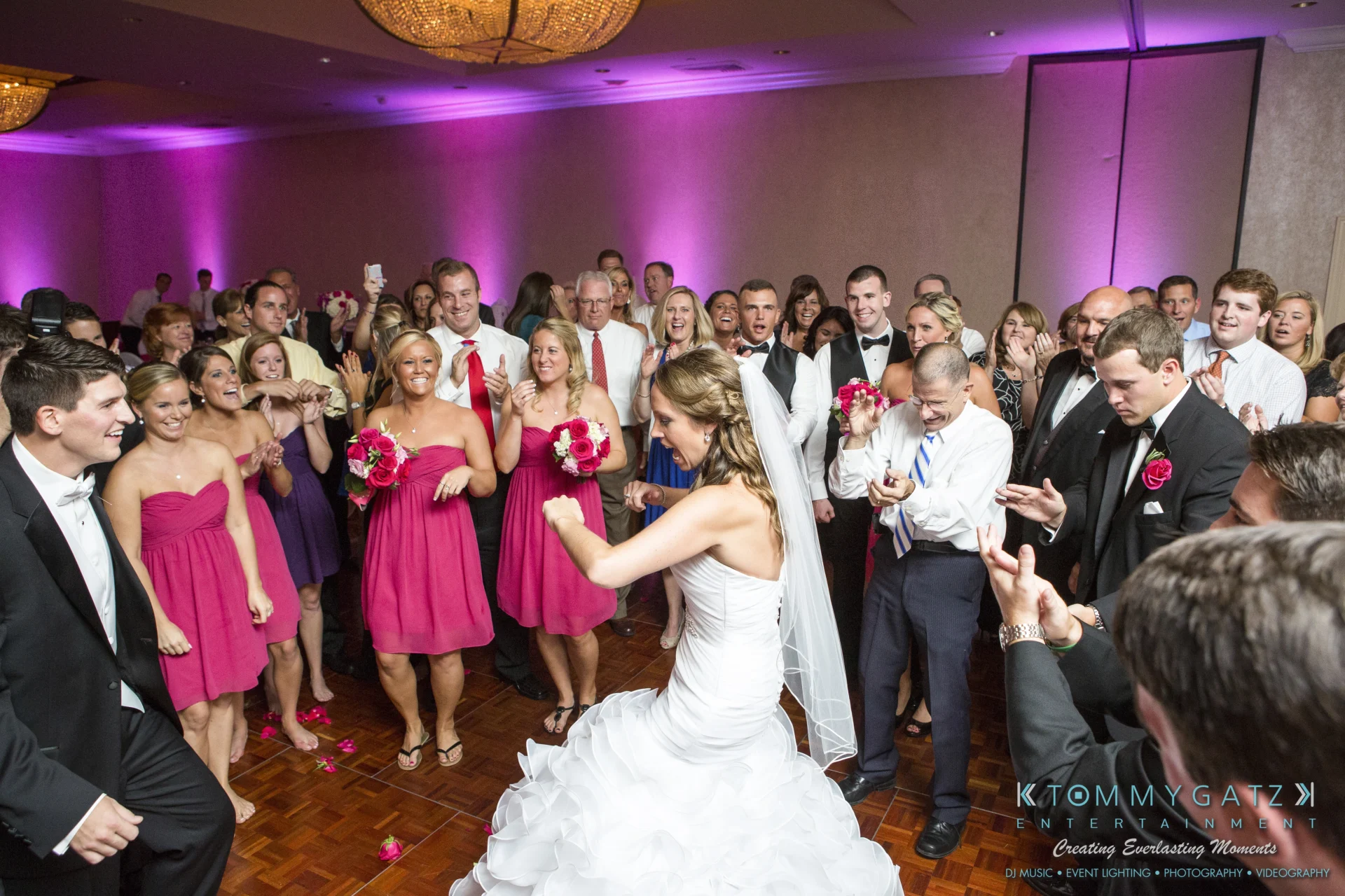 A bride dancing at a wedding reception surrounded by guests. Bridesmaids in pink dresses and groomsmen in black suits are cheering. The room is decorated with pink lighting.