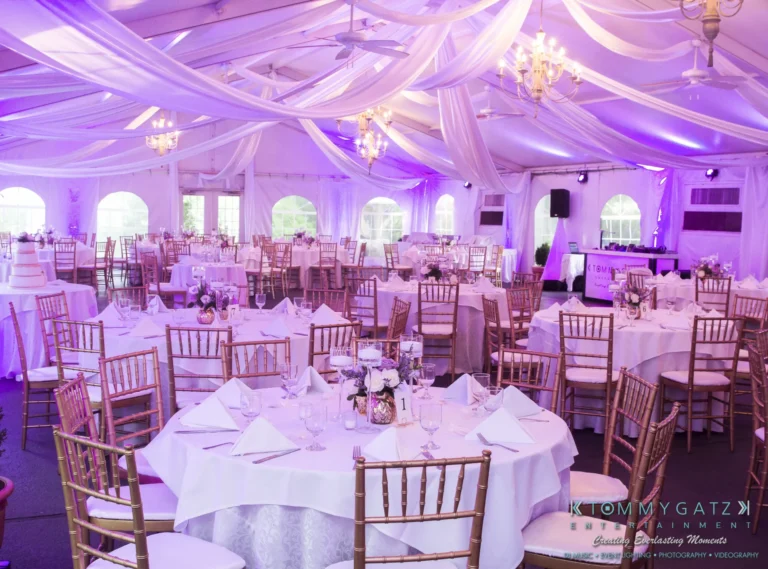 A wedding reception setup in a tent with round tables, white tablecloths, gold chairs, and purple ambient lighting. Tables are decorated with floral centerpieces and place settings.