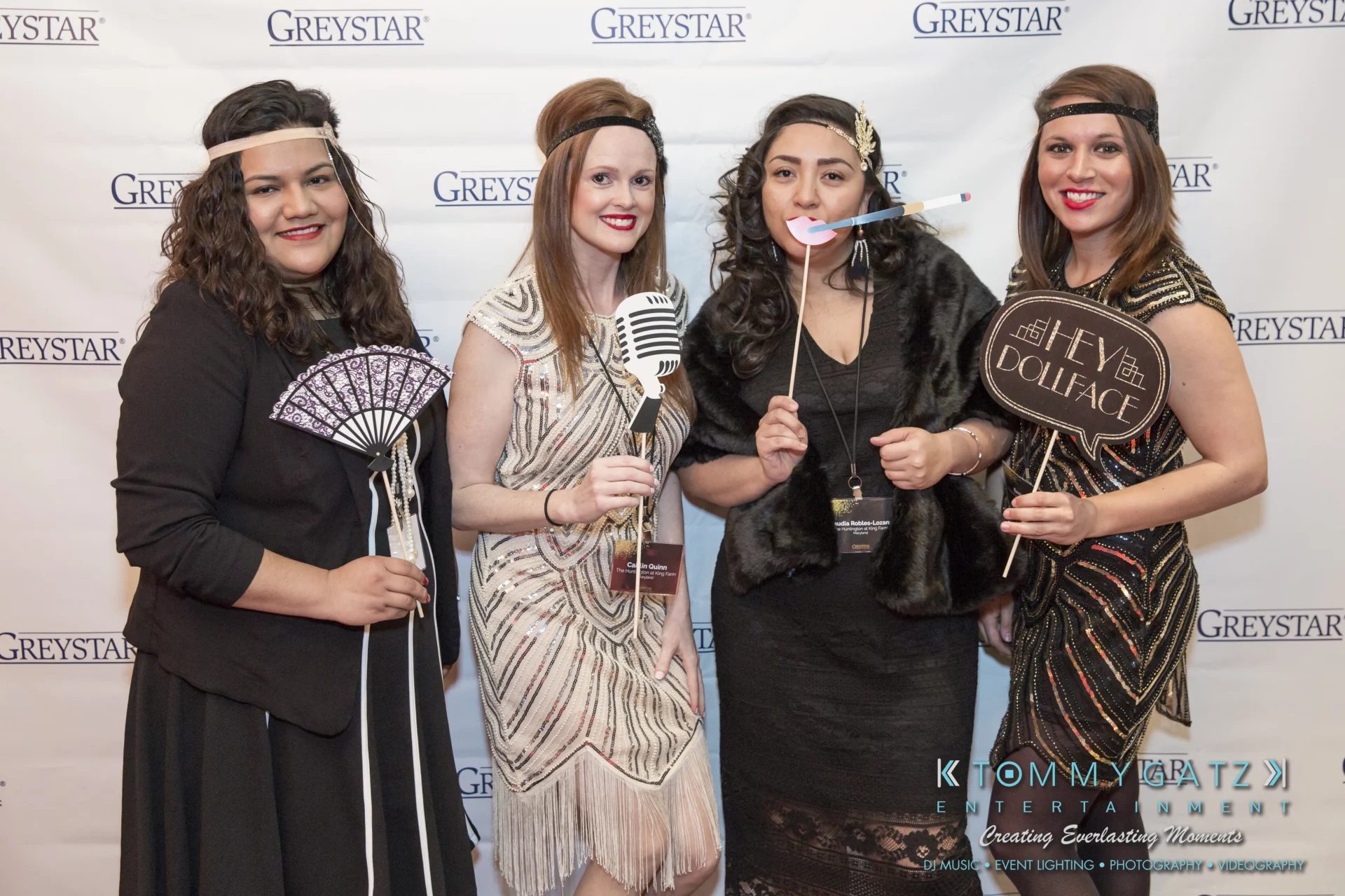 Four women in formal attire stand in front of a backdrop with the word "Greystar." Three hold drinks, and one holds an award labeled "Associate of the Year.