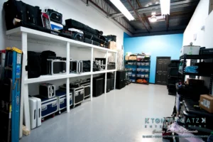 A storage room with organized shelves holding cases, bags, and equipment. A blue accent wall is visible in the background.