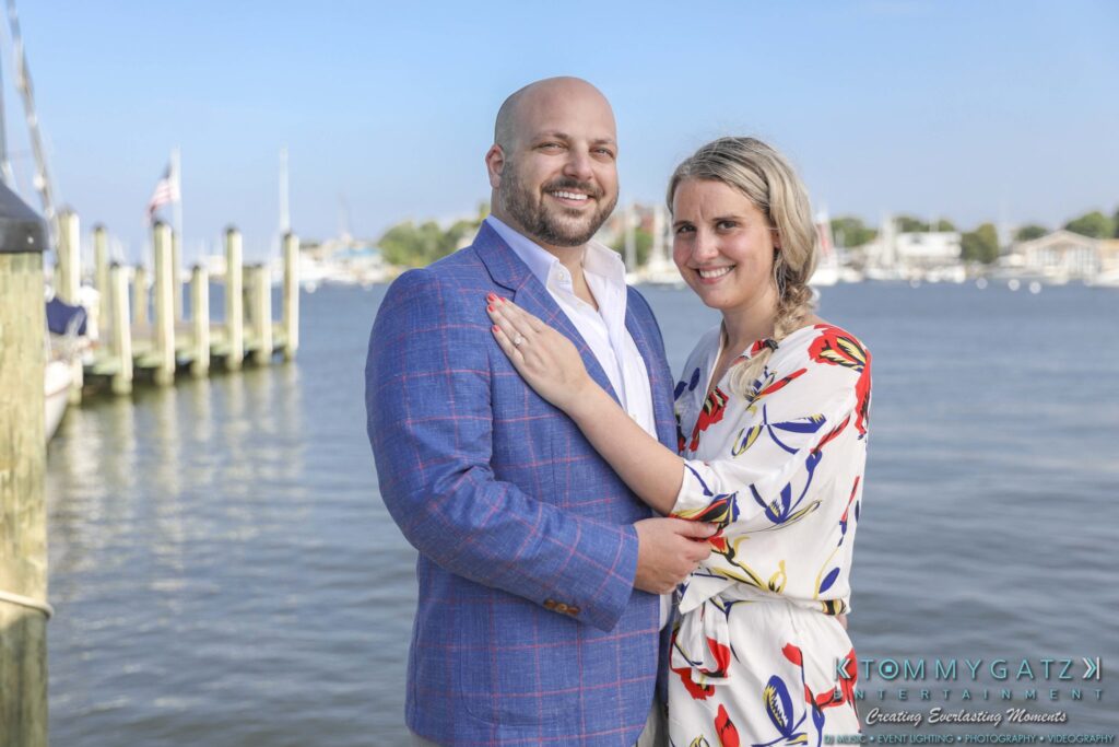 A couple poses together by a waterfront, with boats and docks in the background.