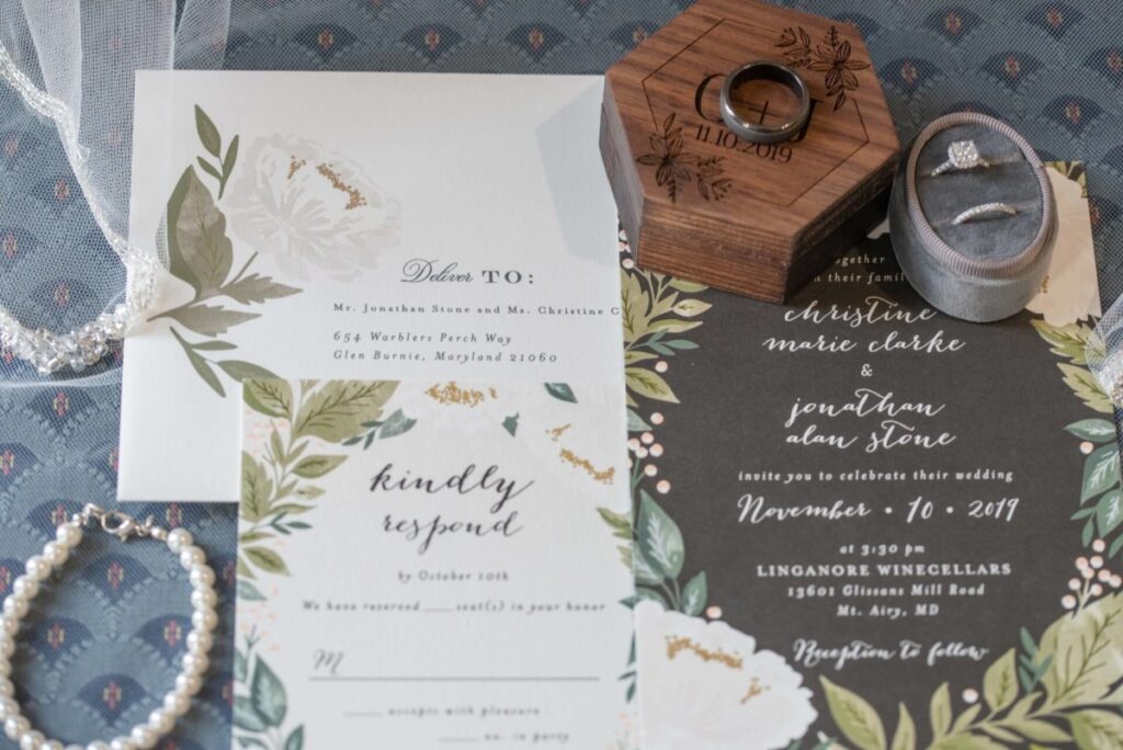 Christine-Jon-Wedding-Blog-1 - TOMMY GATZ ENTERTAINMENT Wedding invitation set with floral designs, including RSVP card, main invitation, and envelope. Features a wooden ring box with engraved date, a ring, and a pearl bracelet on blue fabric.