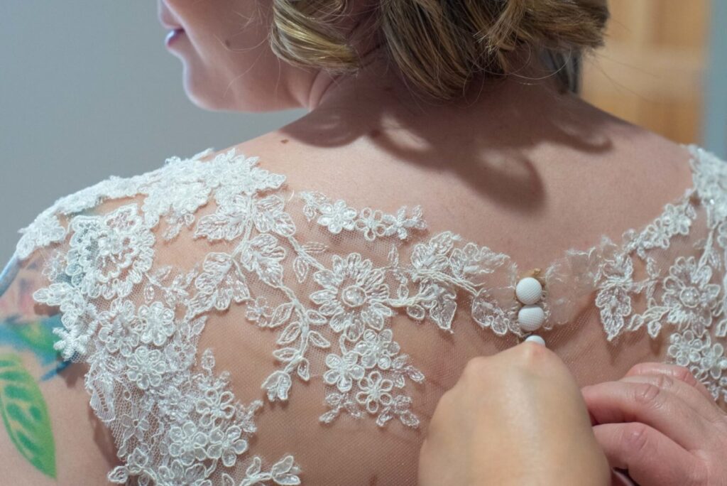 Christine-Jon-Wedding-Blog-10 - TOMMY GATZ ENTERTAINMENT A person fastens buttons on the back of a lace wedding dress.