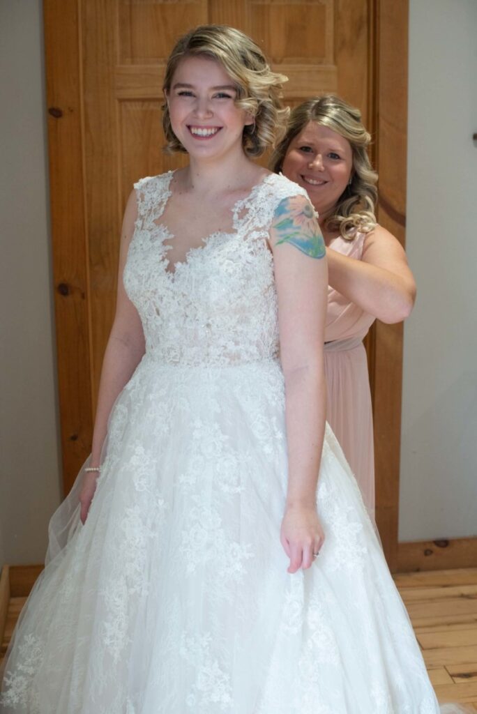Christine-Jon-Wedding-Blog-11 - TOMMY GATZ ENTERTAINMENT A smiling bride in a lace wedding dress stands while another woman in a pink dress helps adjust her gown.