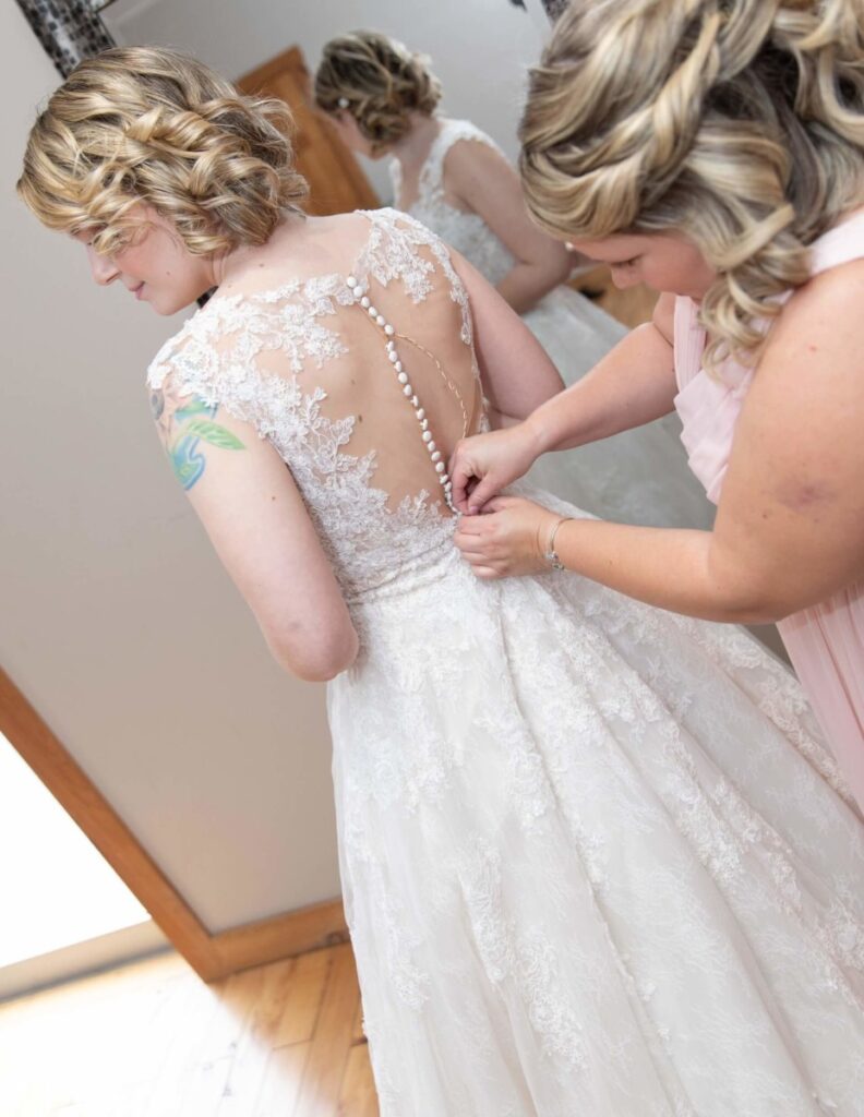 A bride is having her lace wedding dress buttoned by another woman. The dress features an intricate lace back design.