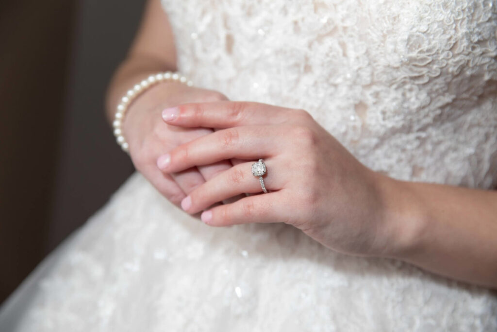 Christine-Jon-Wedding-Blog-13 - TOMMY GATZ ENTERTAINMENT A bride in a lace wedding dress shows her hands clasped together, displaying a pearl bracelet and a ring with a large stone.