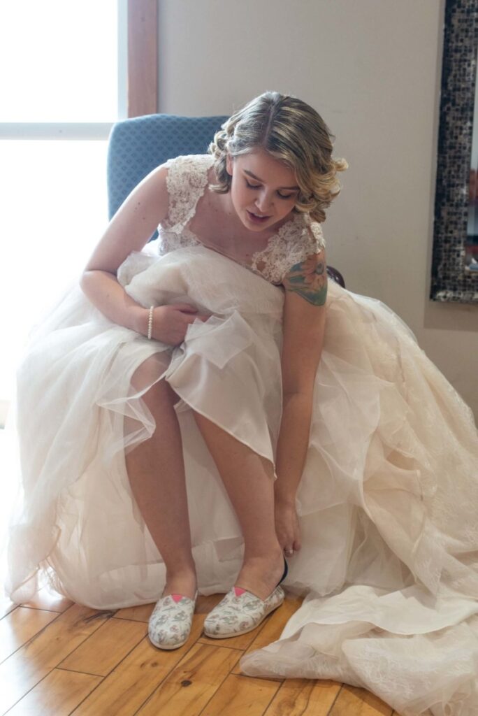 Christine-Jon-Wedding-Blog-14 - TOMMY GATZ ENTERTAINMENT A woman in a white wedding dress adjusts her shoes while seated on a wooden floor.