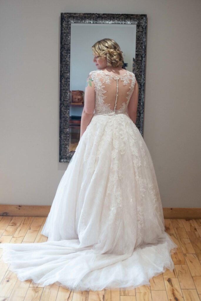 Christine-Jon-Wedding-Blog-15 - TOMMY GATZ ENTERTAINMENT A woman in a lace wedding dress with a long train stands facing a mirror on a wooden floor.