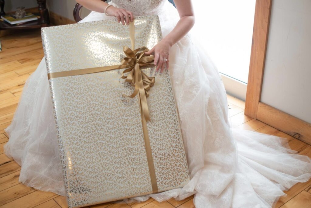 Christine-Jon-Wedding-Blog-16 - TOMMY GATZ ENTERTAINMENT Bride in white gown holding a large, gold gift box with a ribbon, sitting on a wooden floor near a window.
