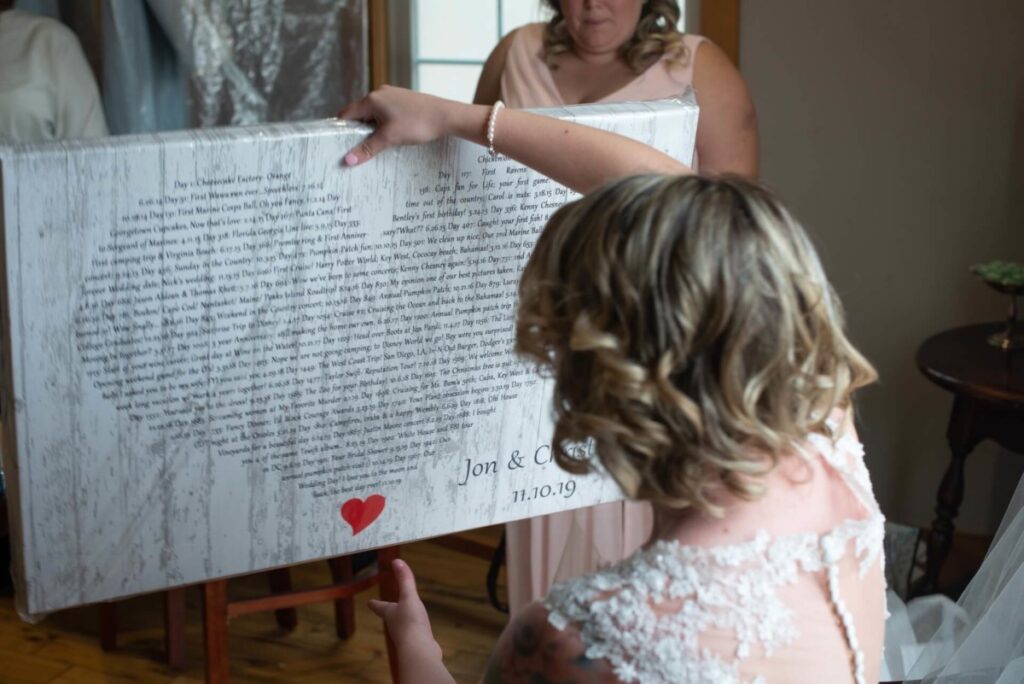 Christine-Jon-Wedding-Blog-17 - TOMMY GATZ ENTERTAINMENT A woman in a wedding dress holds a framed text print with a small red heart and the date 11.10.19 at the bottom. Another woman stands in the background.