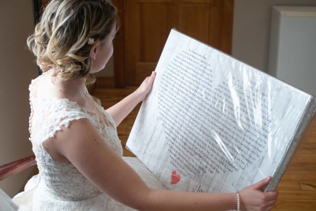 Christine-Jon-Wedding-Blog-18 - TOMMY GATZ ENTERTAINMENT A woman in a lace dress holds a framed artwork featuring text arranged in the shape of a heart.