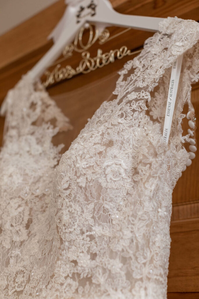 Christine-Jon-Wedding-Blog-2 - TOMMY GATZ ENTERTAINMENT Close-up of a lace wedding dress on a hanger, displaying intricate floral patterns and buttons on the back.