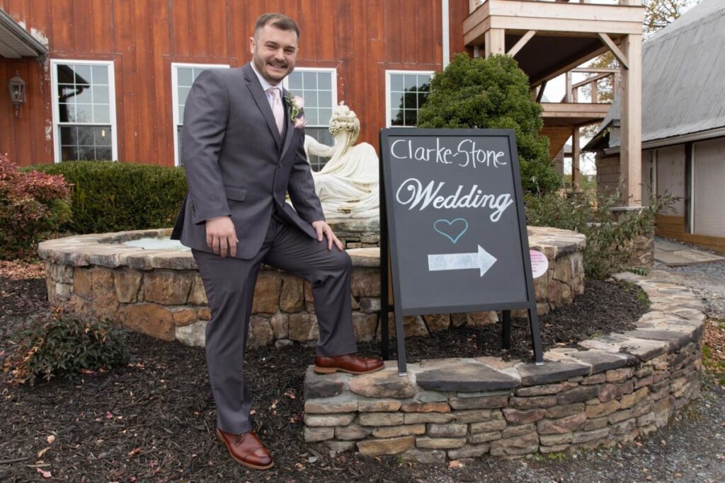 Christine-Jon-Wedding-Blog-20 - TOMMY GATZ ENTERTAINMENT A man in a suit poses by a chalkboard sign reading "Clarke-Stone Wedding" with an arrow. A fountain and rustic building are in the background.