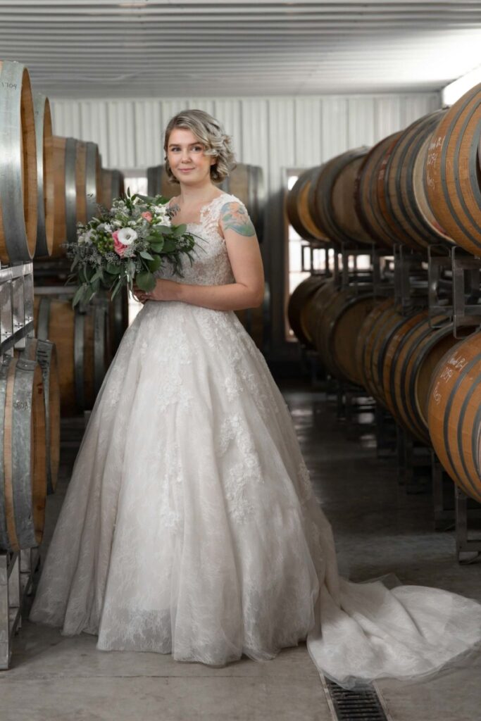 Christine-Jon-Wedding-Blog-21 - TOMMY GATZ ENTERTAINMENT Bride in a lace wedding gown holding a bouquet stands between rows of wine barrels in a cellar.