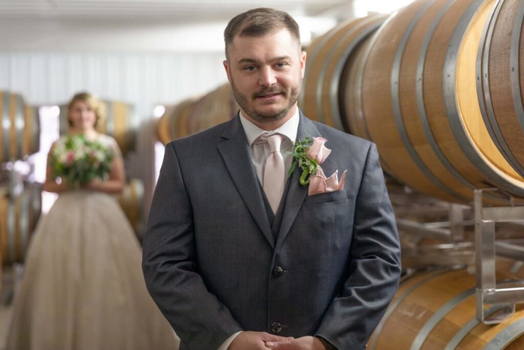 Christine-Jon-Wedding-Blog-22 - TOMMY GATZ ENTERTAINMENT A groom in a suit stands in front of wine barrels, with a bride holding a bouquet in the background.
