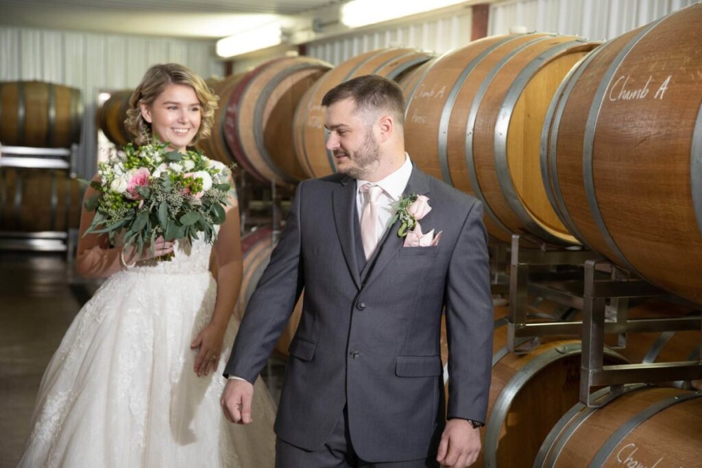 Christine-Jon-Wedding-Blog-23 - TOMMY GATZ ENTERTAINMENT A bride and groom walk past wine barrels, with the groom looking down and the bride holding a bouquet, smiling.
