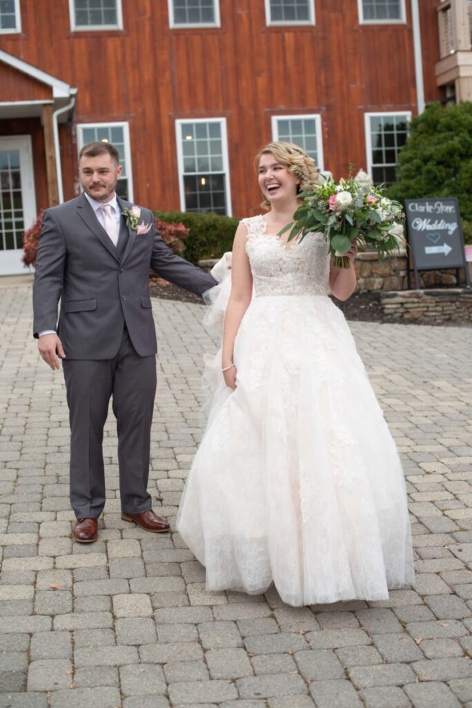 Christine-Jon-Wedding-Blog-24 - TOMMY GATZ ENTERTAINMENT Bride and groom stand outside a building. The bride holds a bouquet and smiles, while the groom stands beside her holding her hand.