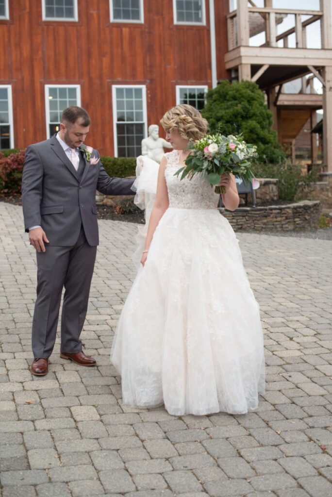 Christine-Jon-Wedding-Blog-25 - TOMMY GATZ ENTERTAINMENT A bride in a white gown and a groom in a gray suit stand outdoors on a cobblestone path, holding hands. The bride holds a bouquet, and a wooden building with windows is in the background.