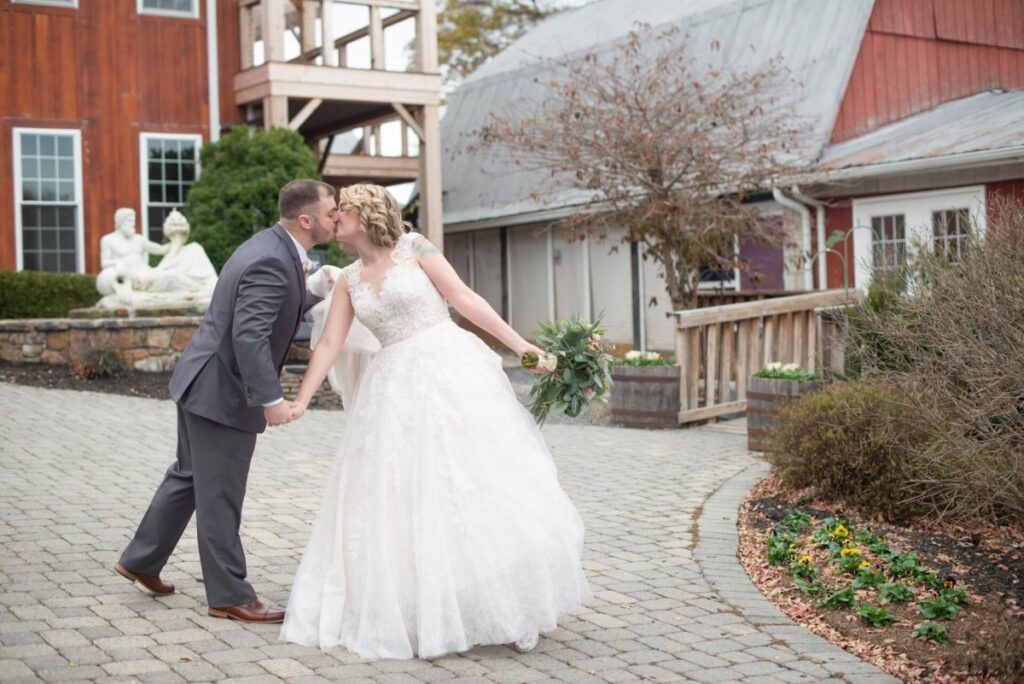 Christine-Jon-Wedding-Blog-26 - TOMMY GATZ ENTERTAINMENT A bride and groom kiss while holding hands on a cobblestone pathway in front of rustic buildings and a small garden. The bride holds a bouquet.