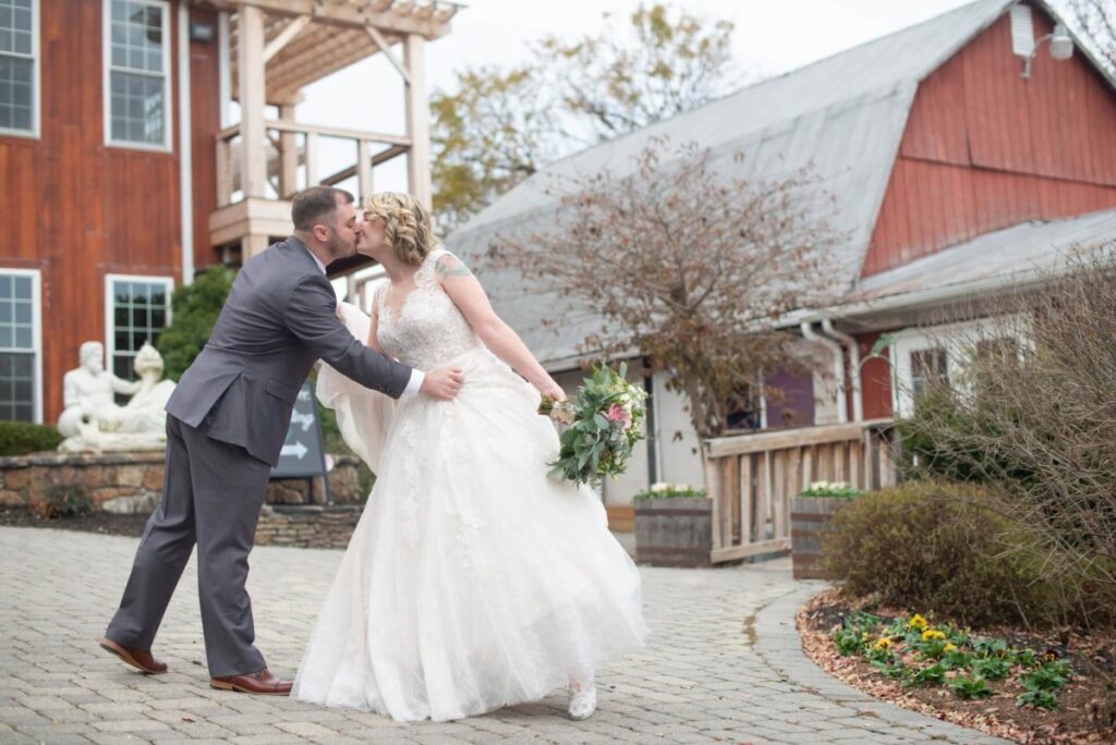 Christine-Jon-Wedding-Blog-27 - TOMMY GATZ ENTERTAINMENT A bride and groom kiss on a cobblestone path in front of a rustic barn and building. The groom is in a gray suit, and the bride wears a white dress, holding a bouquet.