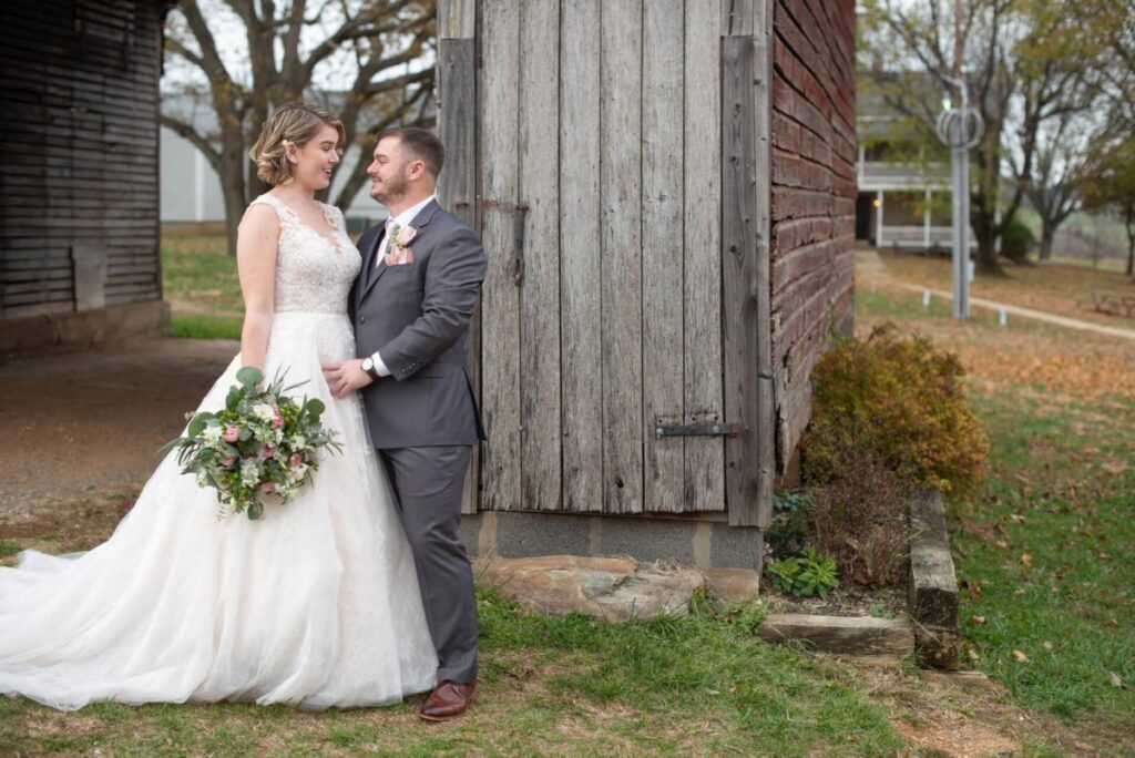 Christine-Jon-Wedding-Blog-28 - TOMMY GATZ ENTERTAINMENT A bride and groom stand together outdoors by a rustic wooden building. The bride holds a bouquet and wears a white dress, and the groom wears a gray suit.