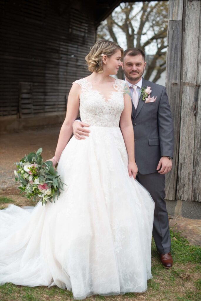 Christine-Jon-Wedding-Blog-29 - TOMMY GATZ ENTERTAINMENT A bride in a white gown holds a bouquet while standing next to a groom in a gray suit with a pink tie. They pose outdoors in front of a wooden structure.
