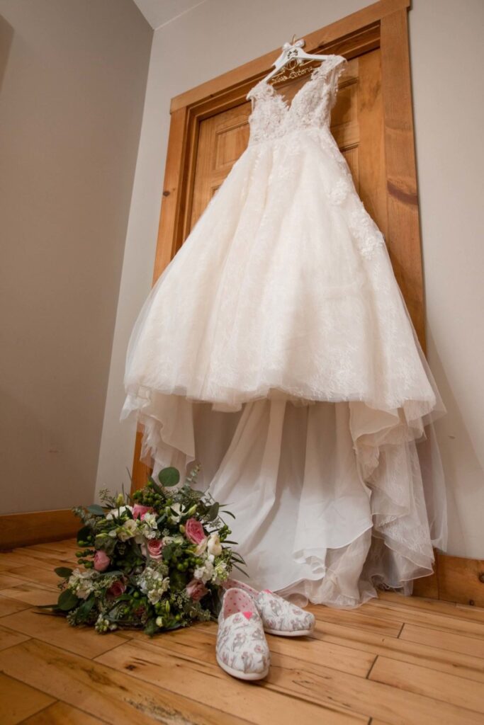 Christine-Jon-Wedding-Blog-3 - TOMMY GATZ ENTERTAINMENT A white wedding dress hangs on a wooden door. Below, there's a floral bouquet and a pair of floral-patterned shoes on a wooden floor.