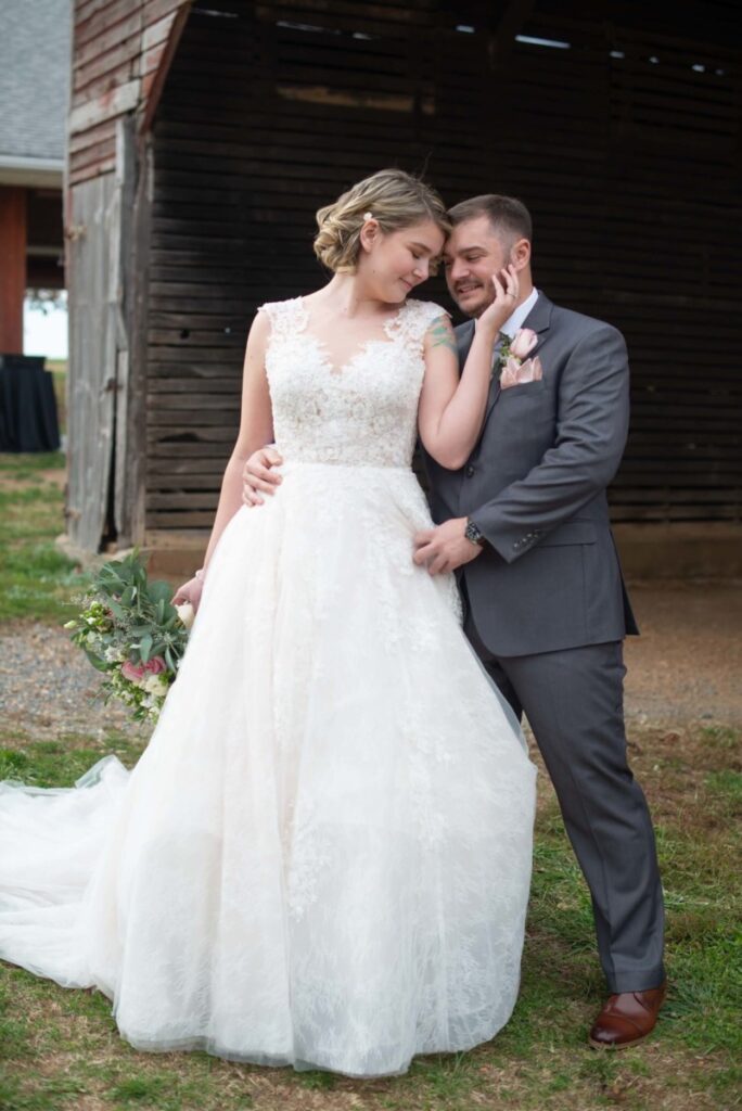 Christine-Jon-Wedding-Blog-30 - TOMMY GATZ ENTERTAINMENT A bride in a white dress and groom in a gray suit pose together outside near a wooden structure. The bride holds a bouquet.