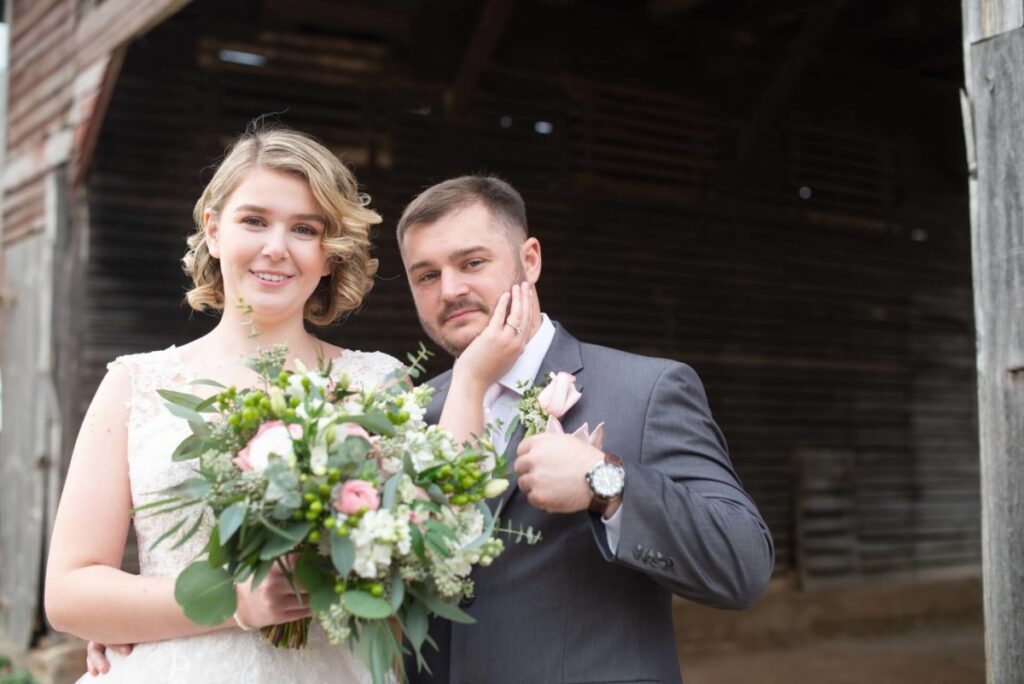 Christine-Jon-Wedding-Blog-31 - TOMMY GATZ ENTERTAINMENT A bride and groom pose for a photo in front of a rustic wooden structure. The bride holds a bouquet of flowers, and the groom touches her face while wearing a suit with a boutonniere.