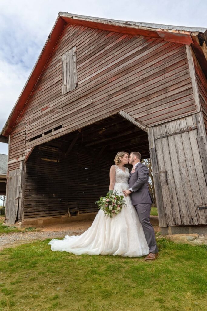 Christine-Jon-Wedding-Blog-32 - TOMMY GATZ ENTERTAINMENT A bride and groom stand together in front of a rustic wooden barn. The bride holds a bouquet, and they gaze at each other.