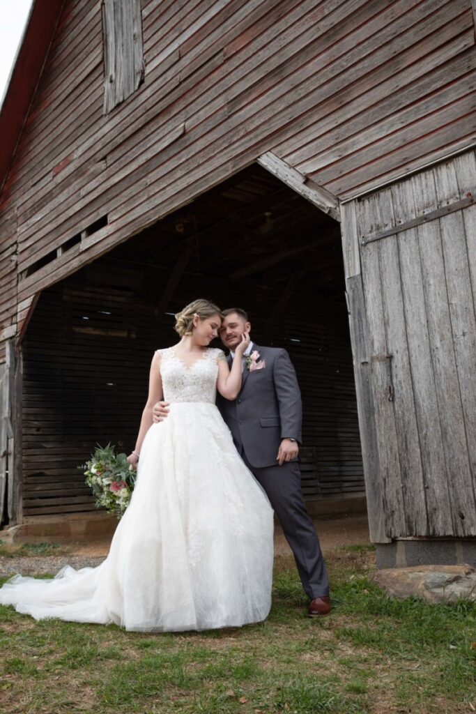 Christine-Jon-Wedding-Blog-33 - TOMMY GATZ ENTERTAINMENT Bride and groom stand affectionately together beside a rustic wooden barn. Bride holds a bouquet, and groom leans against the barn wall.