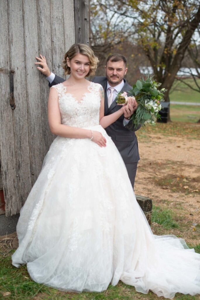 Christine-Jon-Wedding-Blog-34 - TOMMY GATZ ENTERTAINMENT A bride and groom pose outdoors by a wooden structure. The bride holds a bouquet and wears a lace wedding dress, while the groom stands behind her in a suit. Trees are visible in the background.