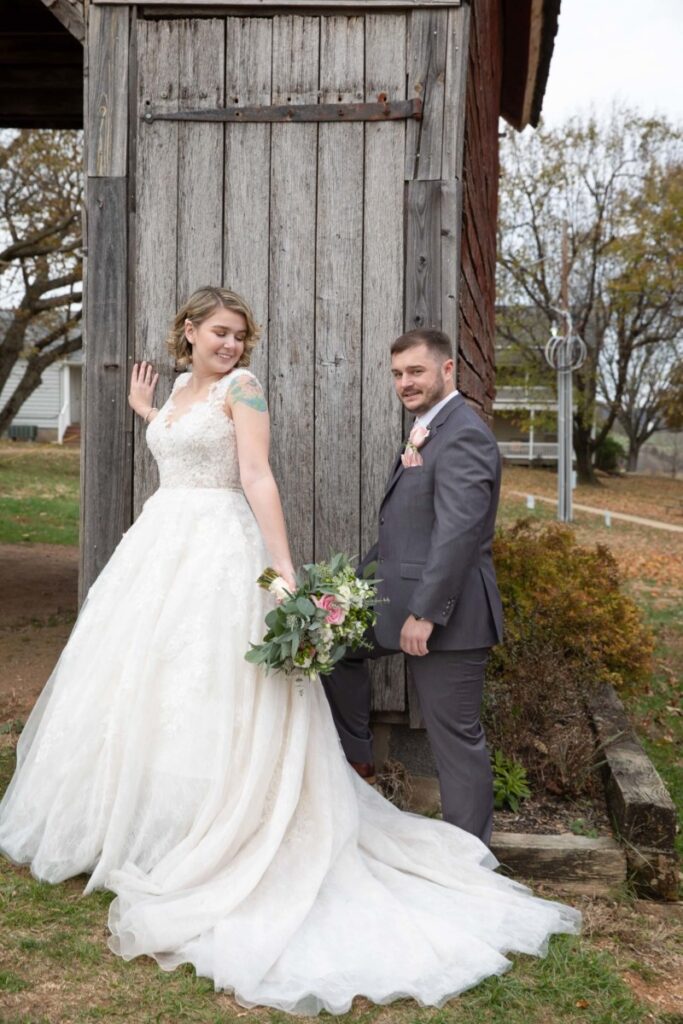 Christine-Jon-Wedding-Blog-35 - TOMMY GATZ ENTERTAINMENT Bride in a white gown and groom in a gray suit stand outside near a wooden structure. The bride holds a bouquet and smiles; the groom has a neutral expression. Trees are visible in the background.