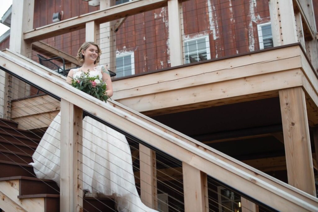 Christine-Jon-Wedding-Blog-36 - TOMMY GATZ ENTERTAINMENT A woman in a white wedding dress holds a bouquet while descending wooden stairs of a rustic building.
