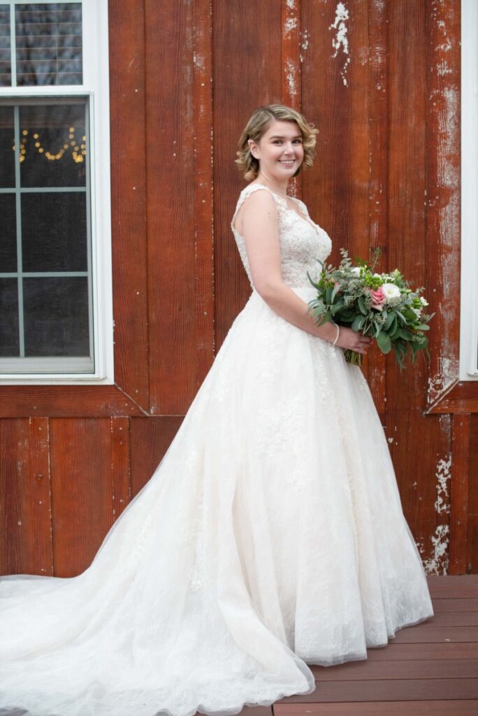 Christine-Jon-Wedding-Blog-37 - TOMMY GATZ ENTERTAINMENT Bride in white lace gown holds a bouquet, standing on a wooden deck against a rustic wooden wall.
