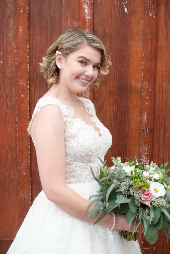 Christine-Jon-Wedding-Blog-38 - TOMMY GATZ ENTERTAINMENT A woman in a white lace dress holding a bouquet, standing against a wooden wall.
