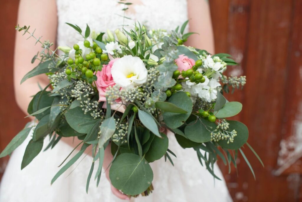 Christine-Jon-Wedding-Blog-39 - TOMMY GATZ ENTERTAINMENT A bride holds a bouquet featuring green leaves, white flowers, pink roses, and small buds, against a textured brown background.