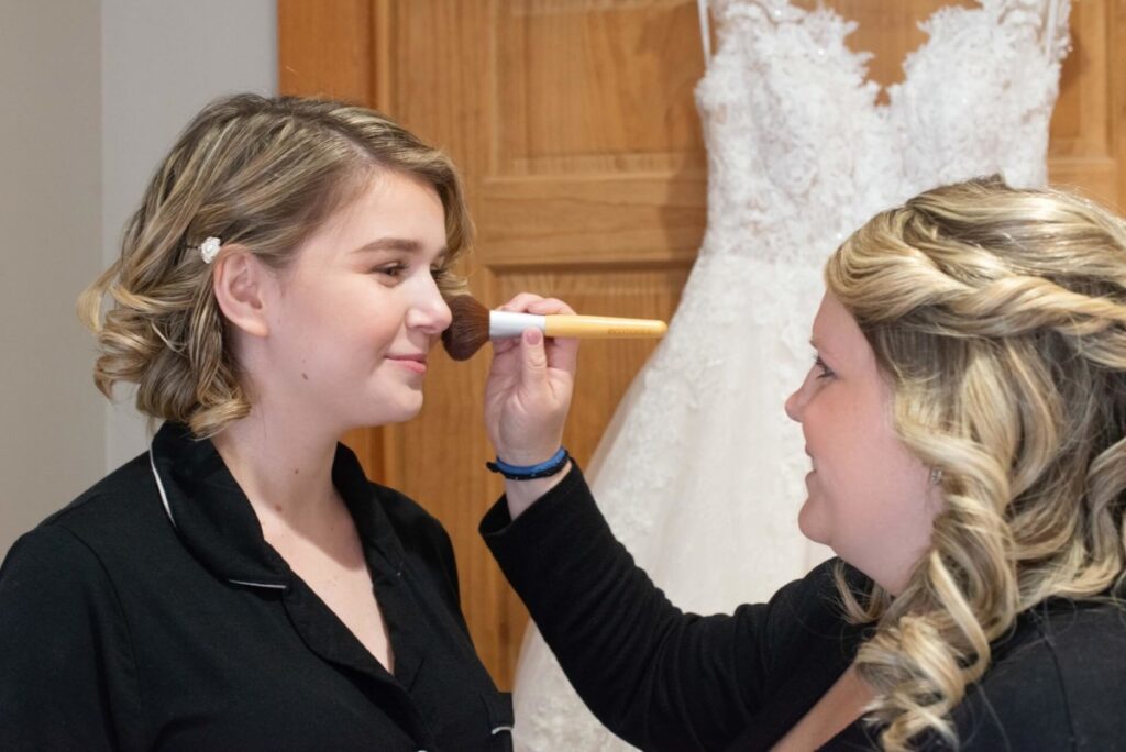 Christine-Jon-Wedding-Blog-4 - TOMMY GATZ ENTERTAINMENT Person applying makeup to another person in front of a white wedding dress hanging on a wooden door.