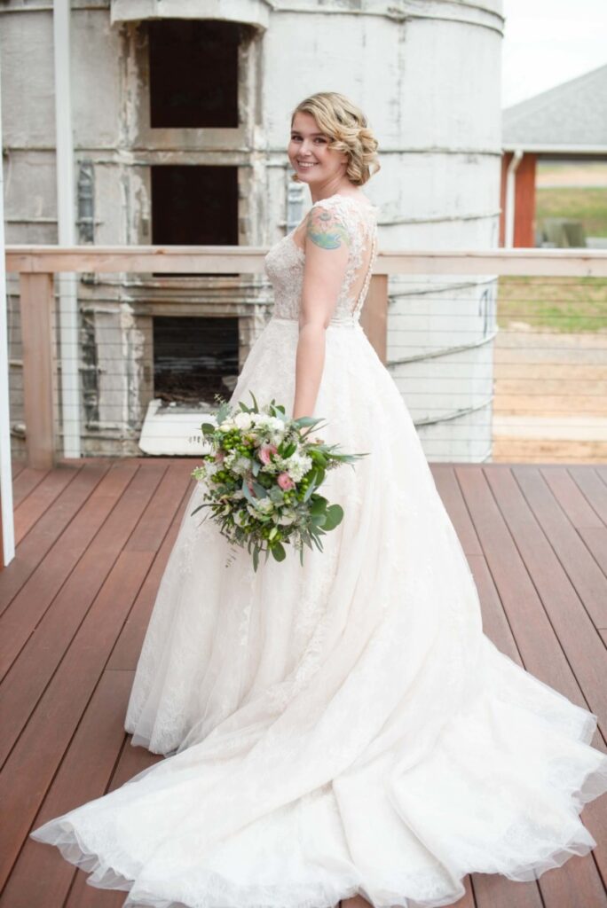 Christine-Jon-Wedding-Blog-40 - TOMMY GATZ ENTERTAINMENT Bride in a white gown holding a bouquet, standing on a wooden deck with a rustic building in the background.