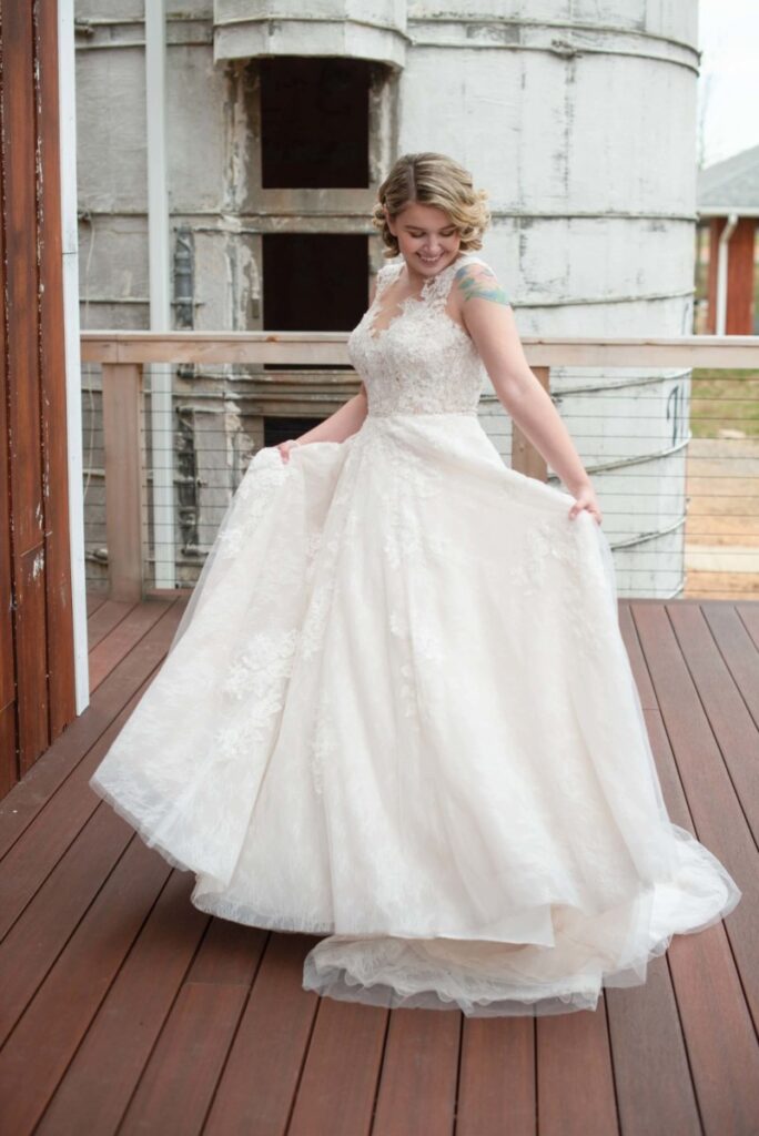 Christine-Jon-Wedding-Blog-41 - TOMMY GATZ ENTERTAINMENT Woman in lace wedding dress smiling and twirling on a wooden deck with large silo in the background.