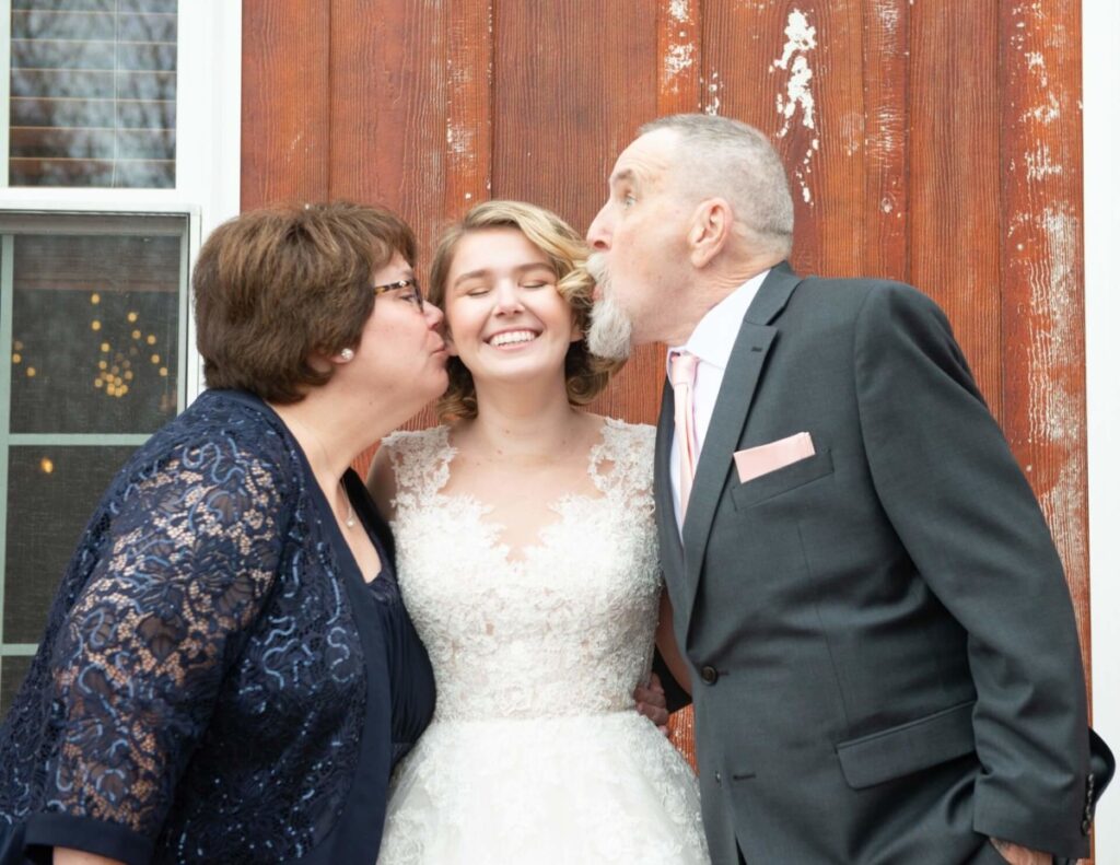 Christine-Jon-Wedding-Blog-42 - TOMMY GATZ ENTERTAINMENT A bride in a lace dress is kissed on both cheeks by a man and woman against a wooden backdrop.