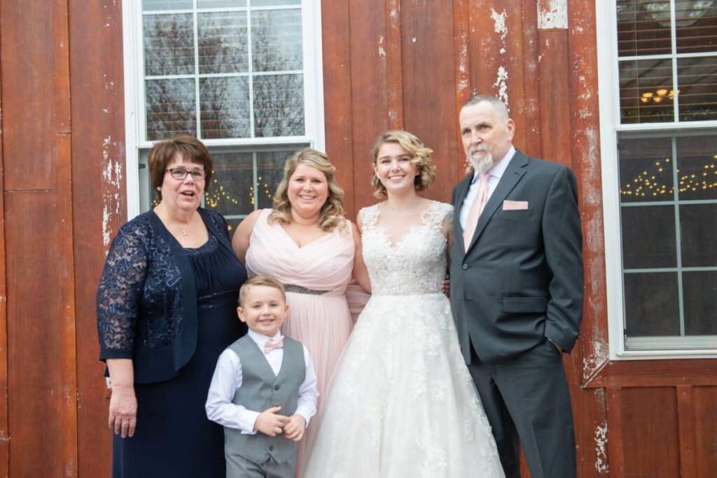 Christine-Jon-Wedding-Blog-43 - TOMMY GATZ ENTERTAINMENT Five people pose for a group photo in formal attire in front of a wooden building with windows.